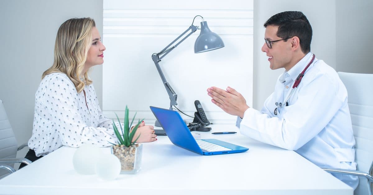 A physician and an adult patient talking across a desk in a bright consulting room